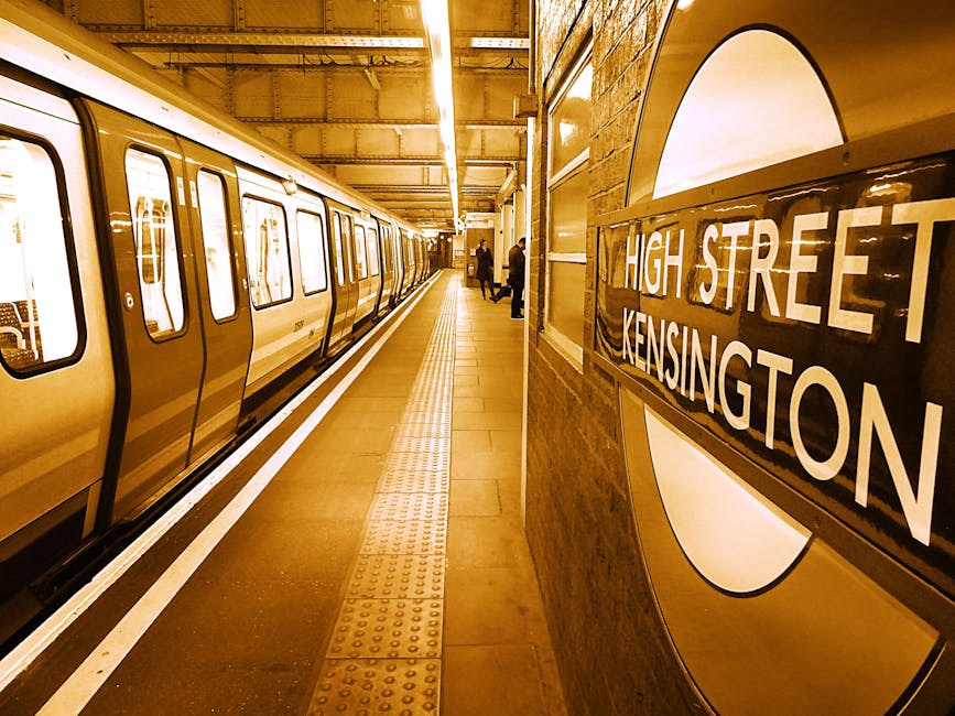 A view of the London Underground platform at High Street Kensington station, showing a yellow-tiled wall with the station sign in white letters on a black background. A train with metallic yellow and